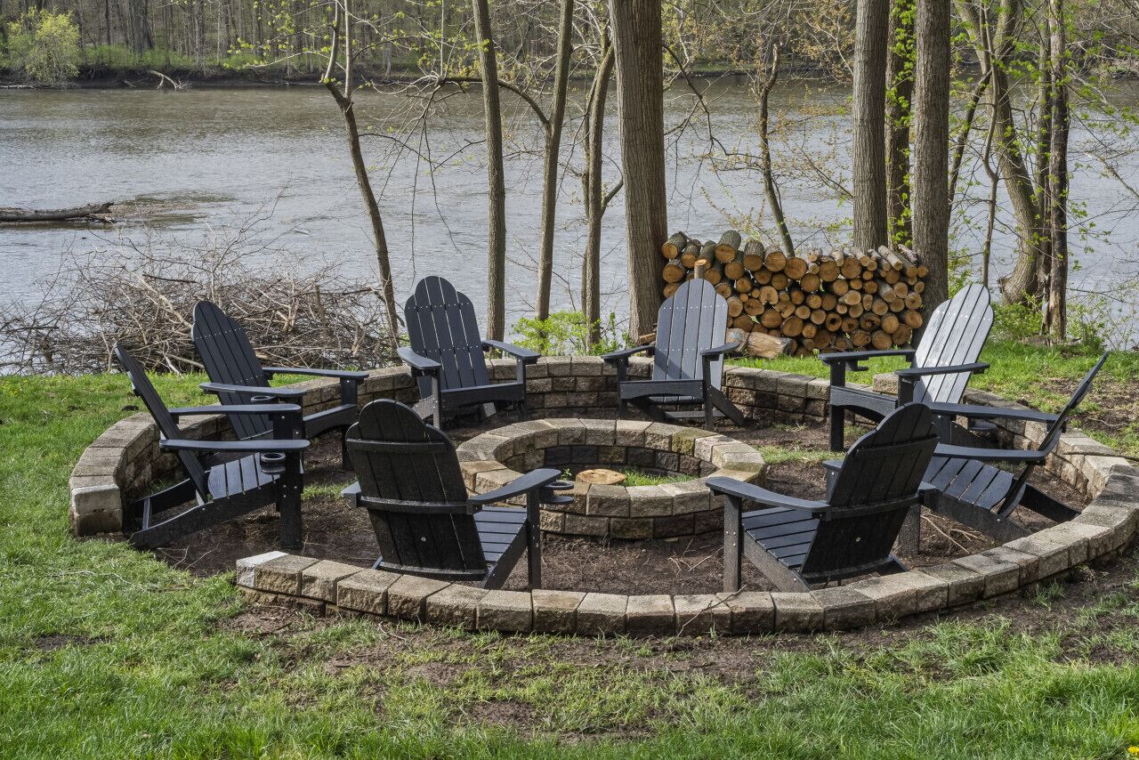 A circular fire pit with Adirondack chairs around it, set in a grassy area with a stack of firewood and a lake in the background.