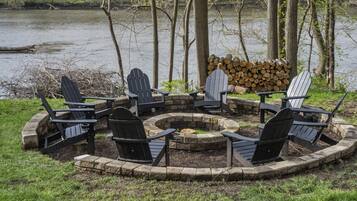 A circular fire pit with Adirondack chairs around it, set in a grassy area with a stack of firewood and a lake in the background.