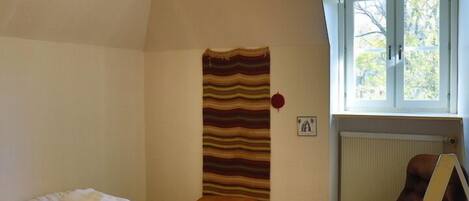 A room with a bed, a chair, a desk with papers, a radiator, a window with a view of trees, and a hanging tapestry.