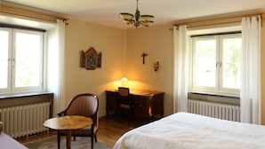 A room with a bed, a desk with a chair, a small table, and a radiator. There are two windows with curtains, a cross on the wall, and a chandelier. - Birgittasystrarnas Gästhem (Vadstena)