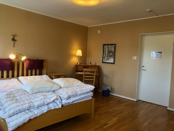 A bedroom with a wooden bed, a nightstand, a desk, a chair, a lamp, a framed picture, a door, and a trash bin. - Birgittasystrarnas Gästhem (Vadstena)