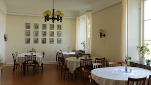 A dining area with multiple round tables and chairs, a chandelier, framed pictures on the wall, and potted plants. - Birgittasystrarnas Gästhem (Vadstena)