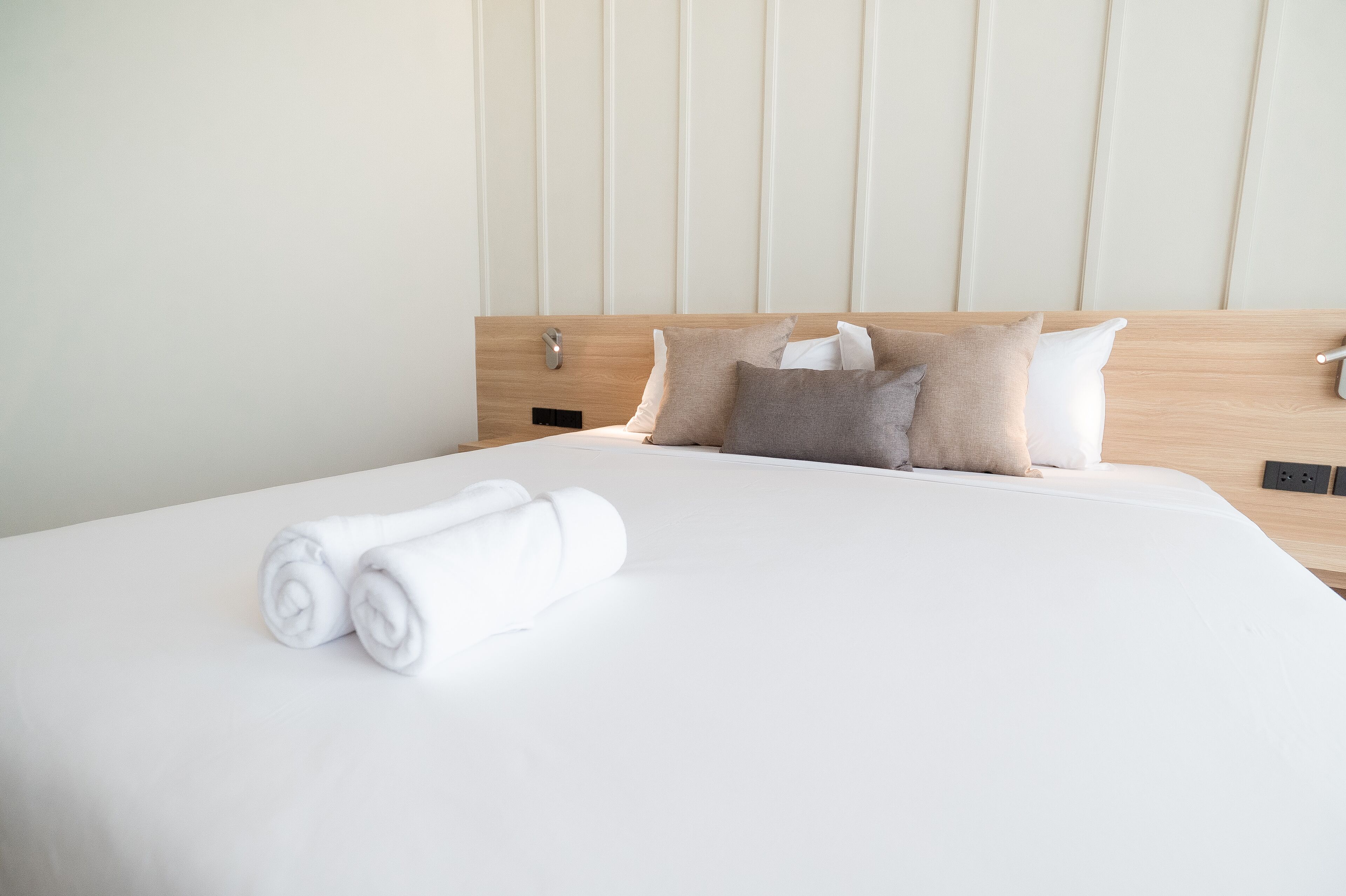 Ein ordentlich gemachtes Bett mit weißen Leinen und aufgerollten Handtüchern, einem Holz-Kopfstück und einer Wand in neutralen Farbtönen.