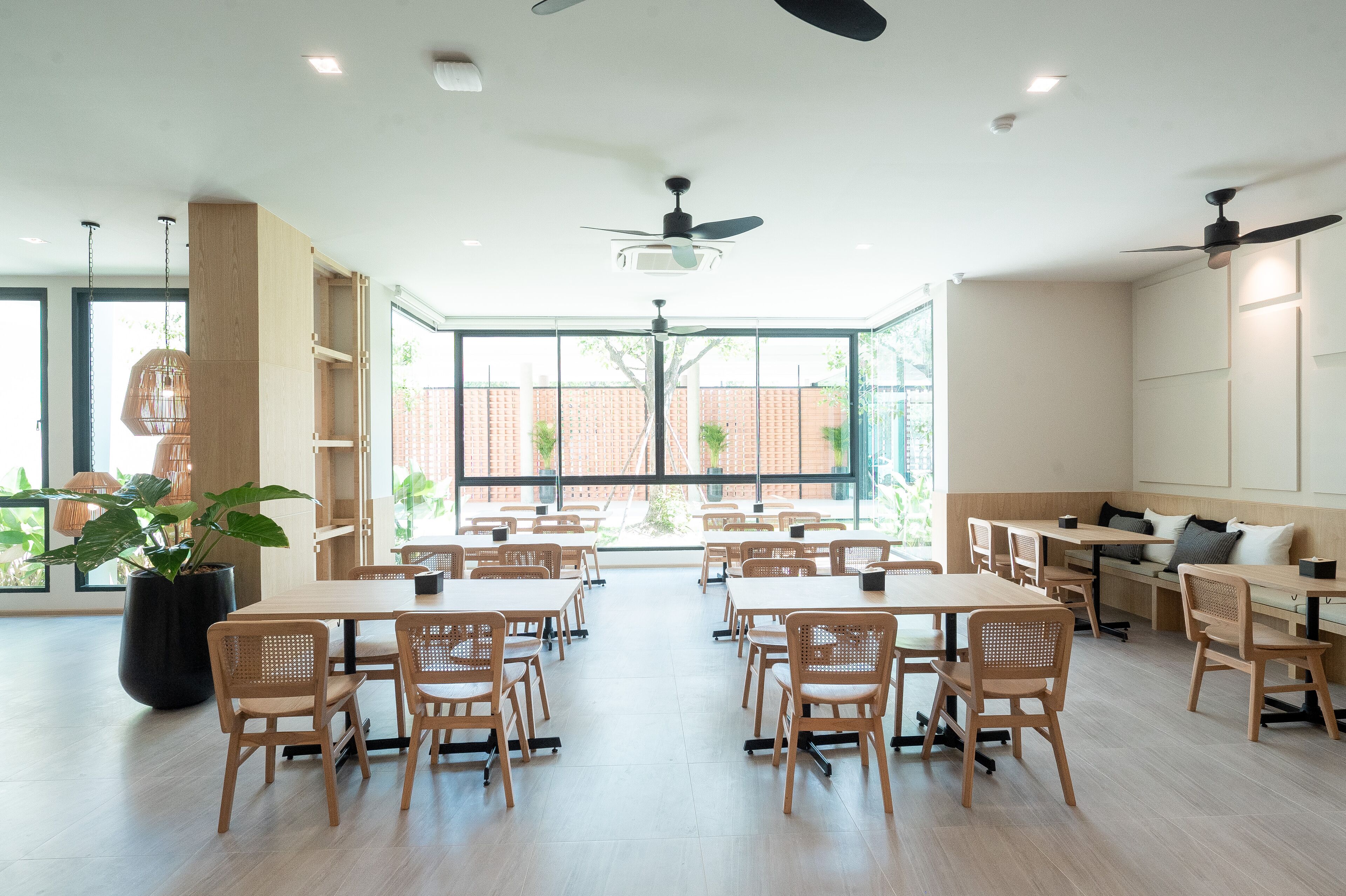 A spacious dining area with wooden tables and chairs, large windows with a view of a brick building, and ceiling fans.