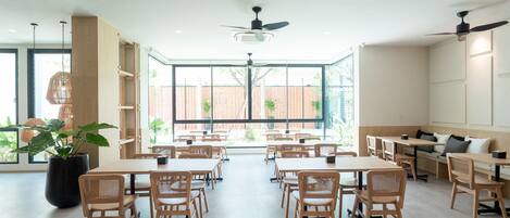 A spacious dining area with wooden tables and chairs, large windows with a view of a brick building, and ceiling fans.