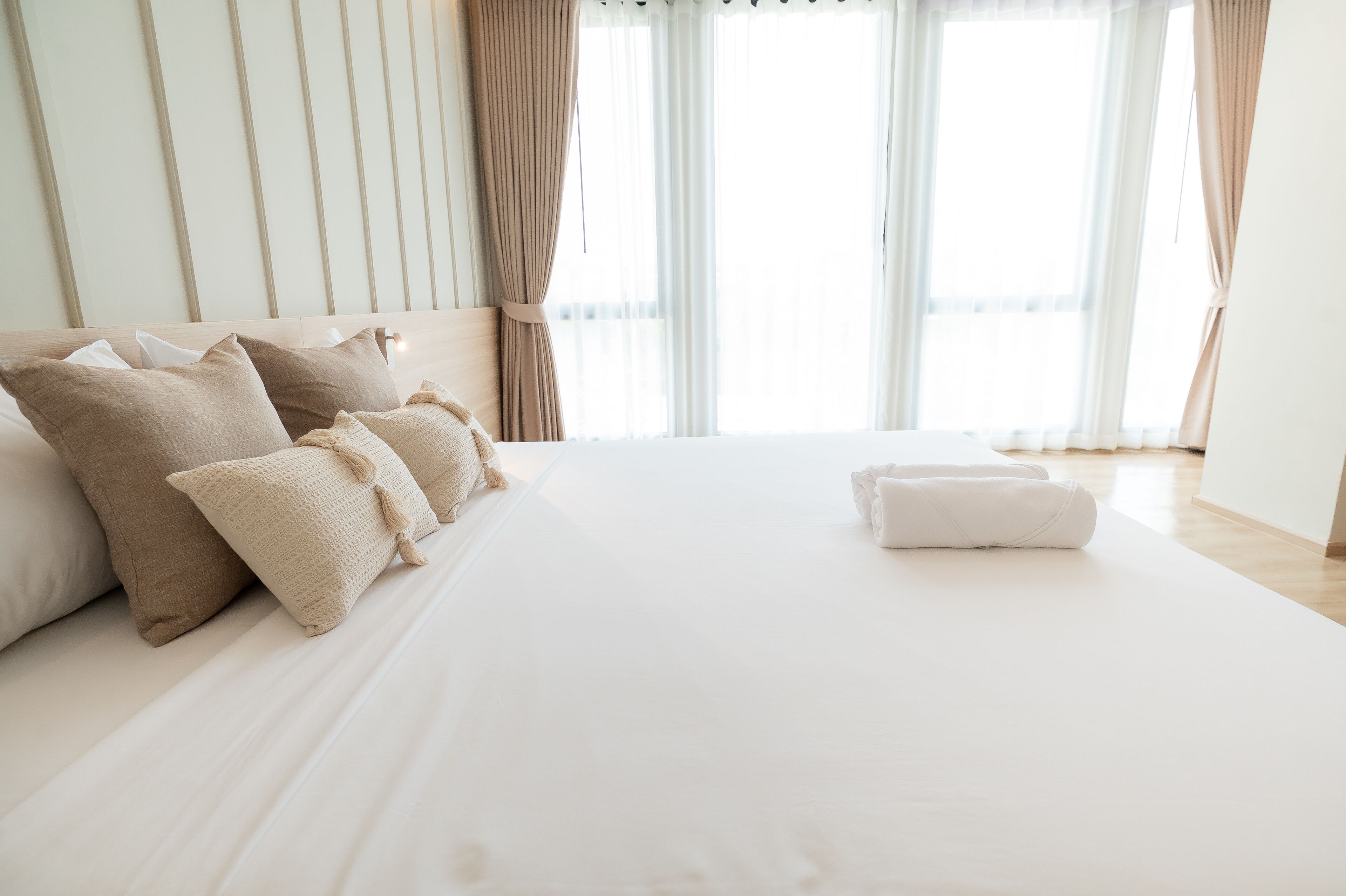 A neatly made bed with beige and white bedding, a white pillow, and a rolled-up towel on a wooden floor.