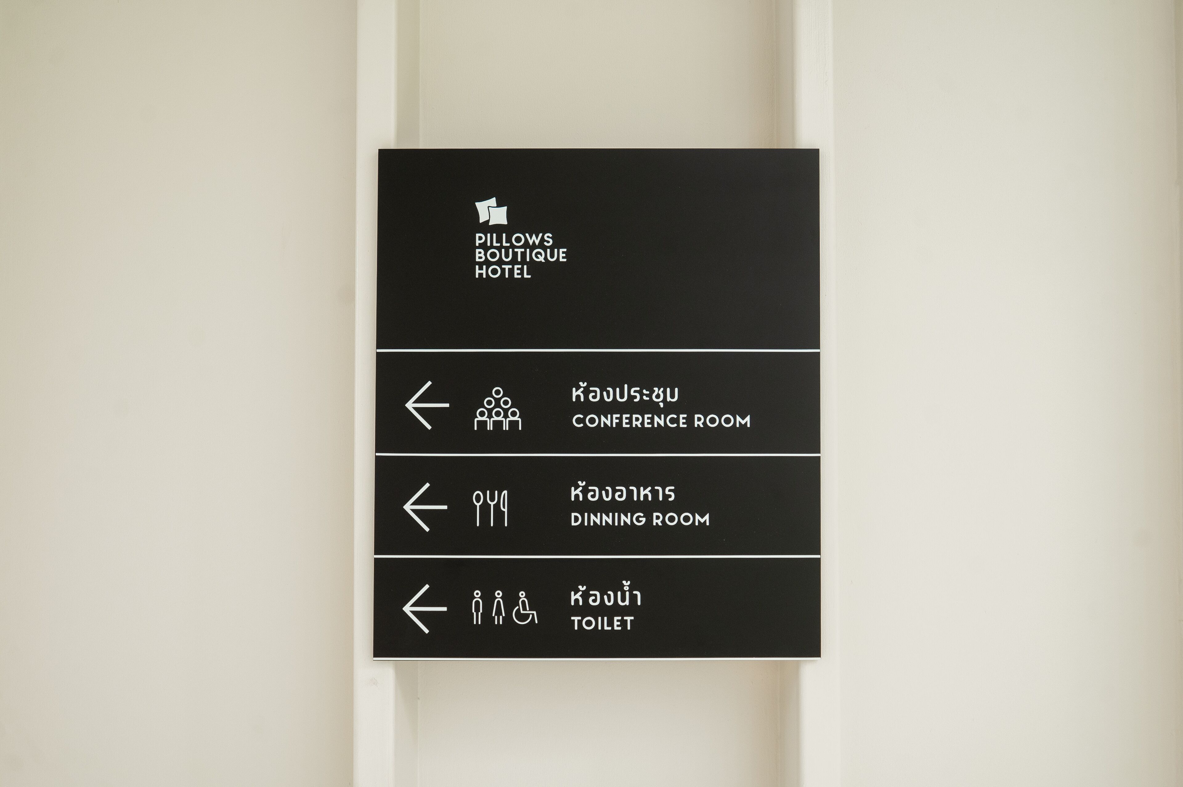 Señal negra con texto e iconos blancos que indican la ubicación de la sala de conferencias, el comedor y el baño en el hotel boutique Pillows.