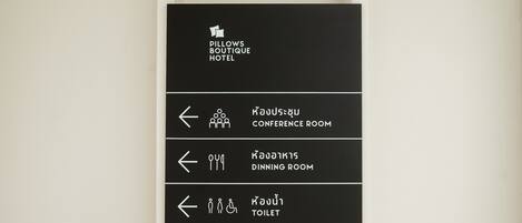 Señal negra con texto e iconos blancos que indican la ubicación de la sala de conferencias, el comedor y el baño en el hotel boutique Pillows.