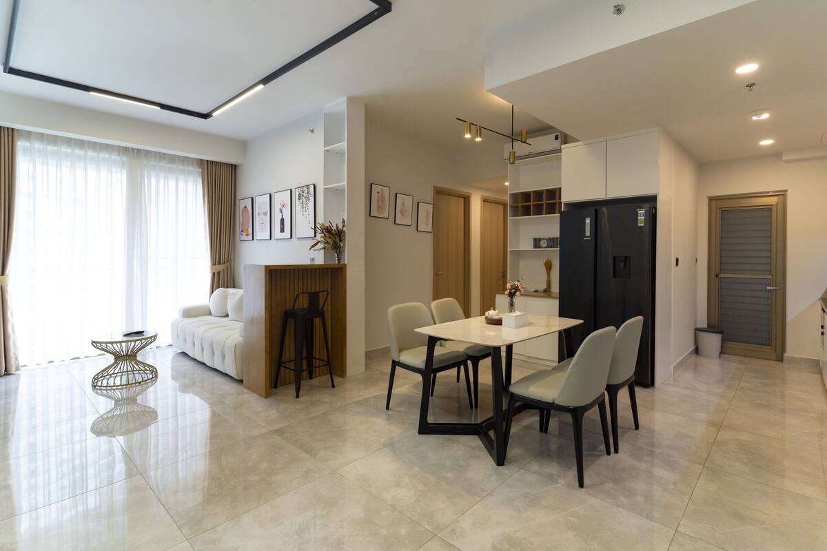 Ein modernes Wohnzimmer mit einer Couch, einem Esstisch und Stühlen. Es gibt eine Küchenzeile mit Kühlschrank und Schränken.