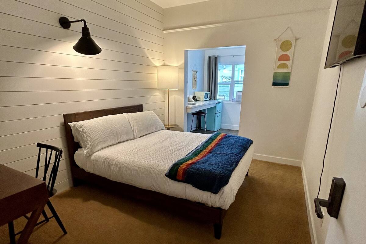 Ein Schlafzimmer mit einem Bett, einem Schreibtisch und einem Stuhl. Ein Fenster bietet Blick ins Freie.