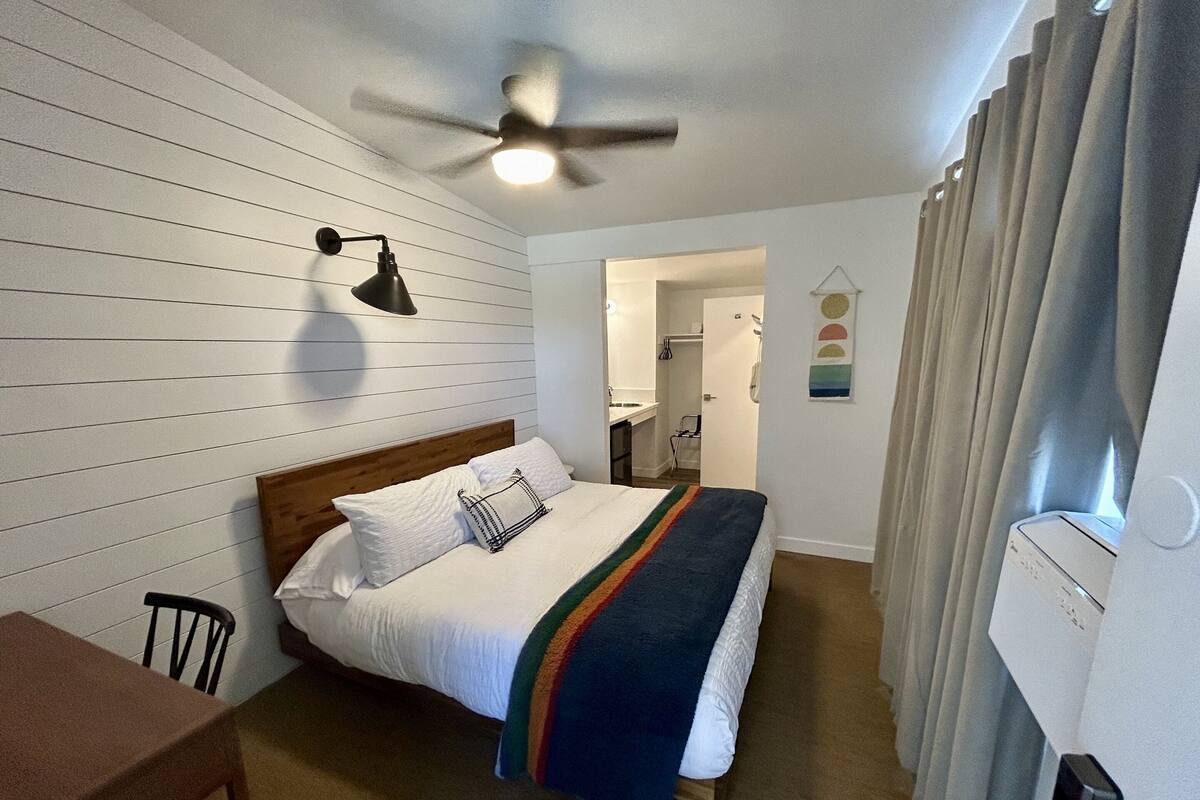 Ein Schlafzimmer mit Bett, Schreibtisch, Deckenventilator und einem durchgegangenen Türrahmen sichtbaren Badezimmer.