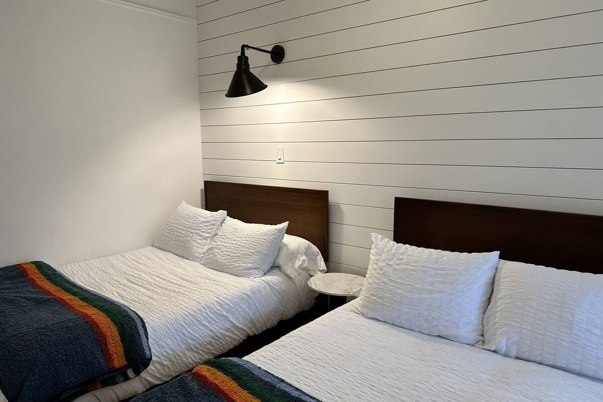 Ein Schlafzimmer mit zwei Betten, weißer Bettwäsche, einem Holzkopfende, einem Nachttisch und einer Wandleuchte.