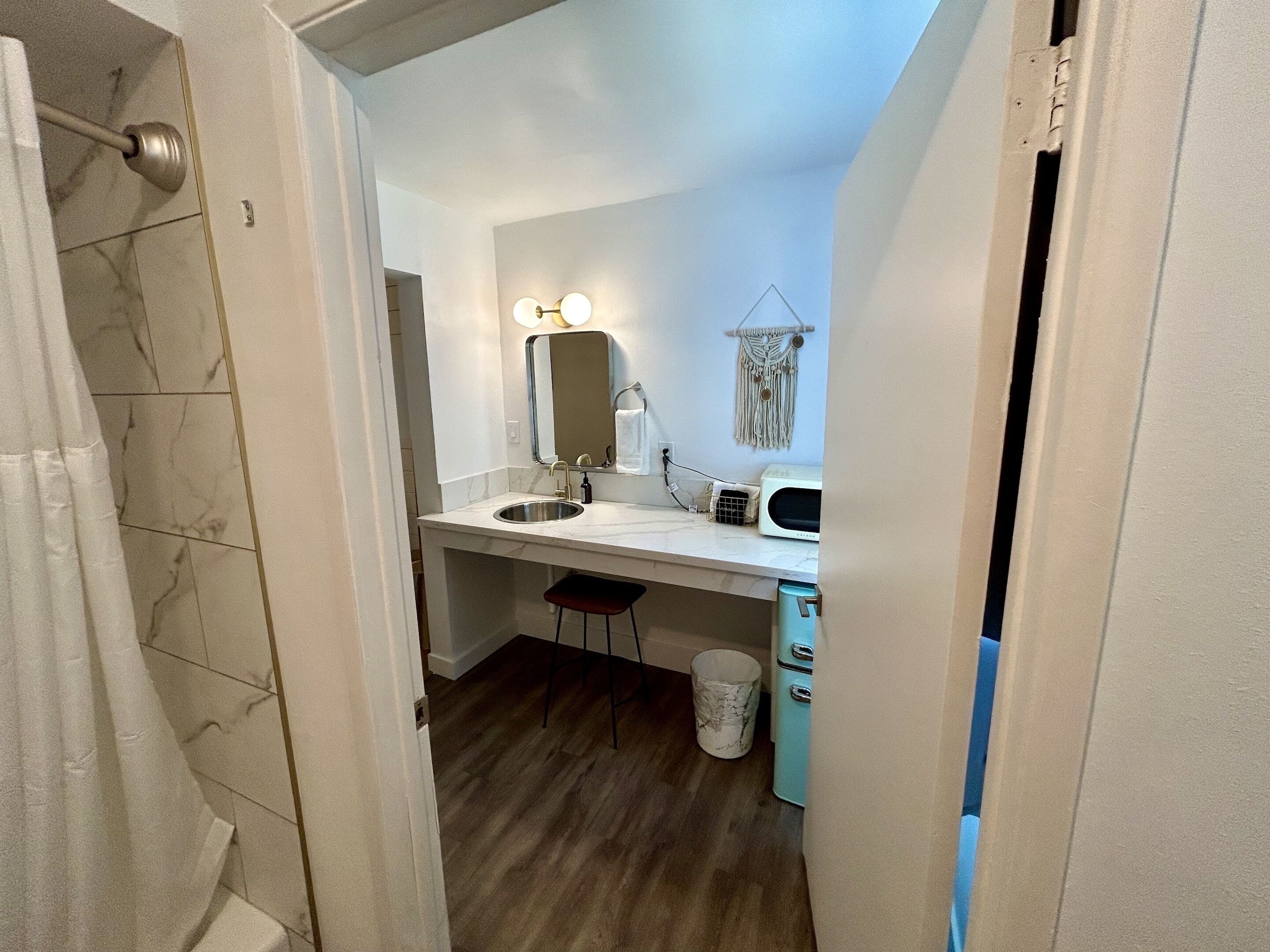 Un baño con lavamanos de mármol, un espejo con iluminación, un perchero, un taburete pequeño y un cubo de basura.