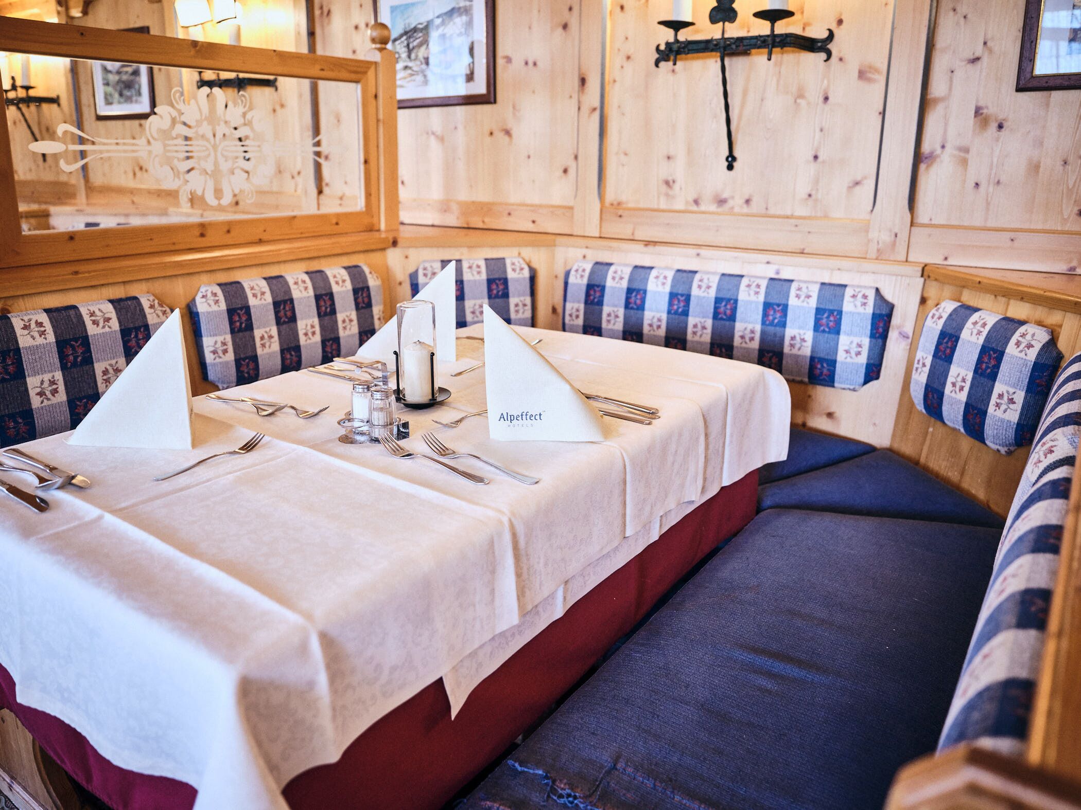 A dining table set with a white tablecloth, silverware, and a candle centerpiece. The room features wooden walls, checkered cushions, and a mirror.