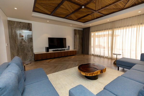 Una sala de estar moderna con un televisor de pantalla plana grande, sofás azules, una mesa de centro de madera y un techo con motivo de espiga.