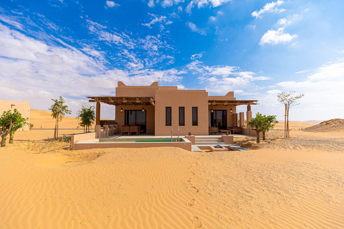 Une maison située dans le désert, dotée d’un patio, d’une piscine et offrant une vue sur les dunes environnantes.