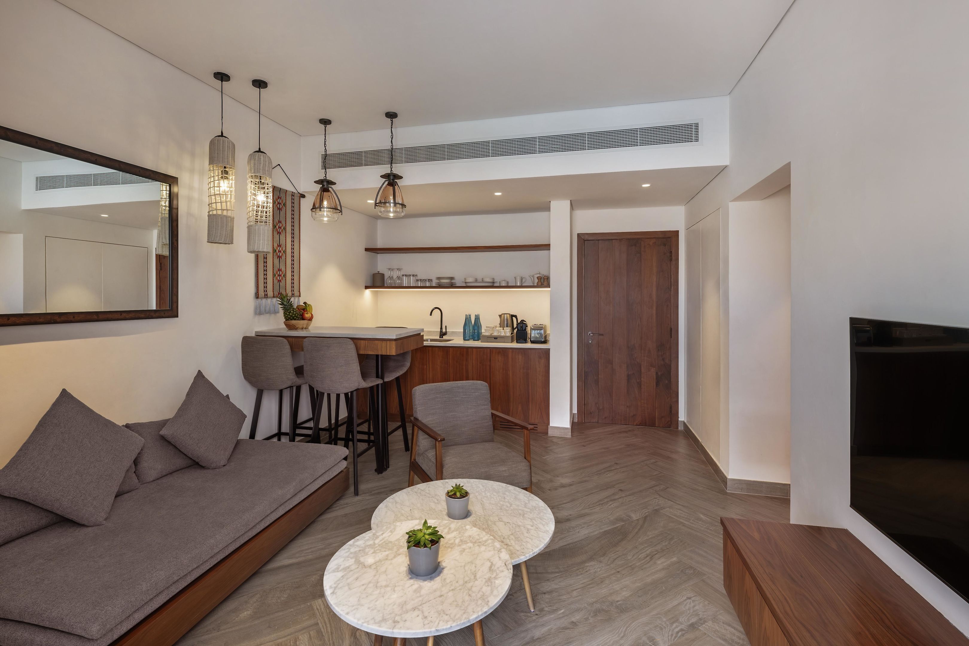 Een moderne woonkamer met een bank, twee ronde tafels met plantenpotten, een keuken met barkrukken, en een grote televisie.