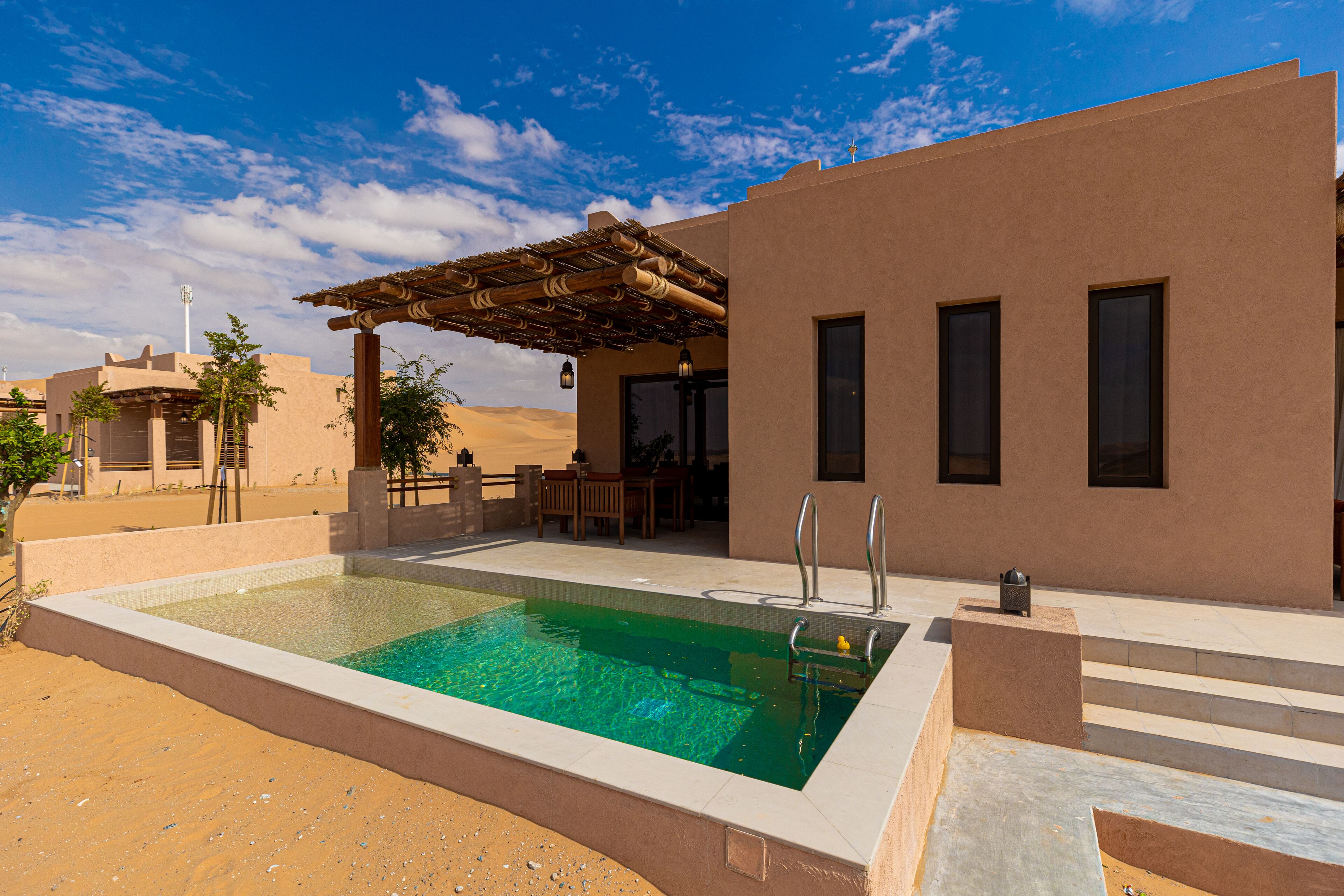 A swimming pool with a pergola, a covered patio, and a desert landscape in the background.