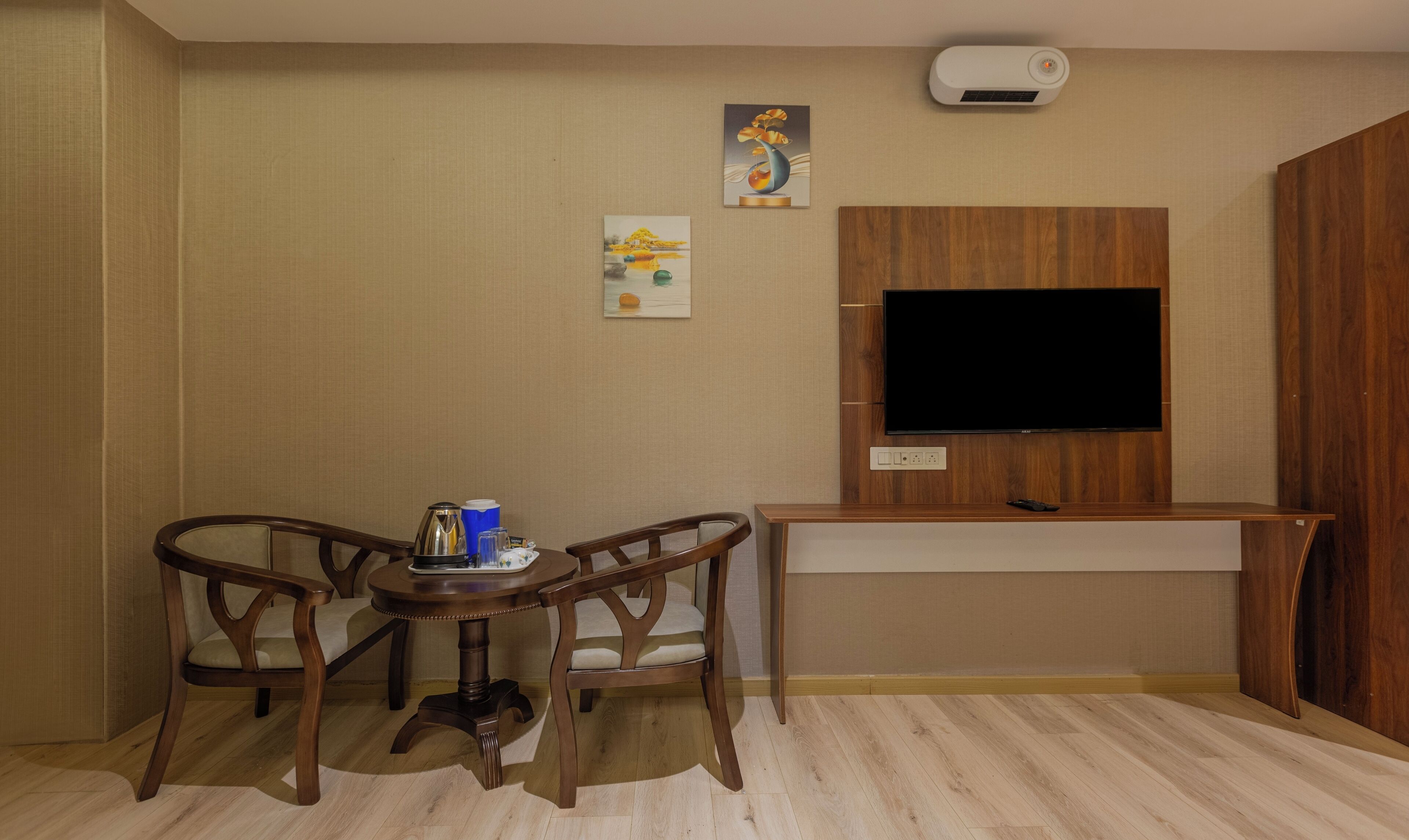 A small, neatly arranged room with a wooden desk, two chairs, a flat-screen TV mounted on a wooden panel, and a wall-mounted air conditioner.
