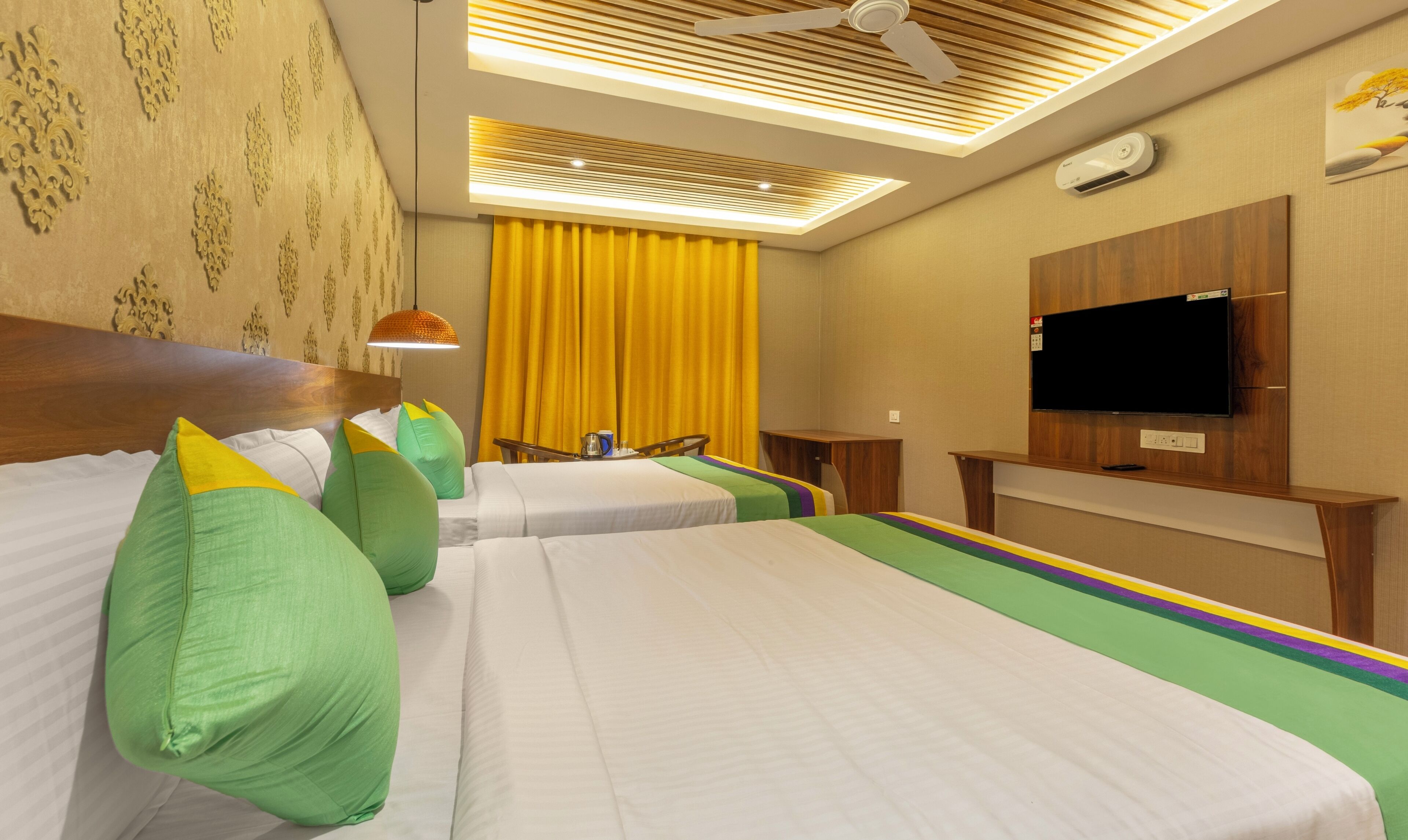 Habitación de hotel con dos camas, televisión, ventilador de techo y ventana con cortinas amarillas.