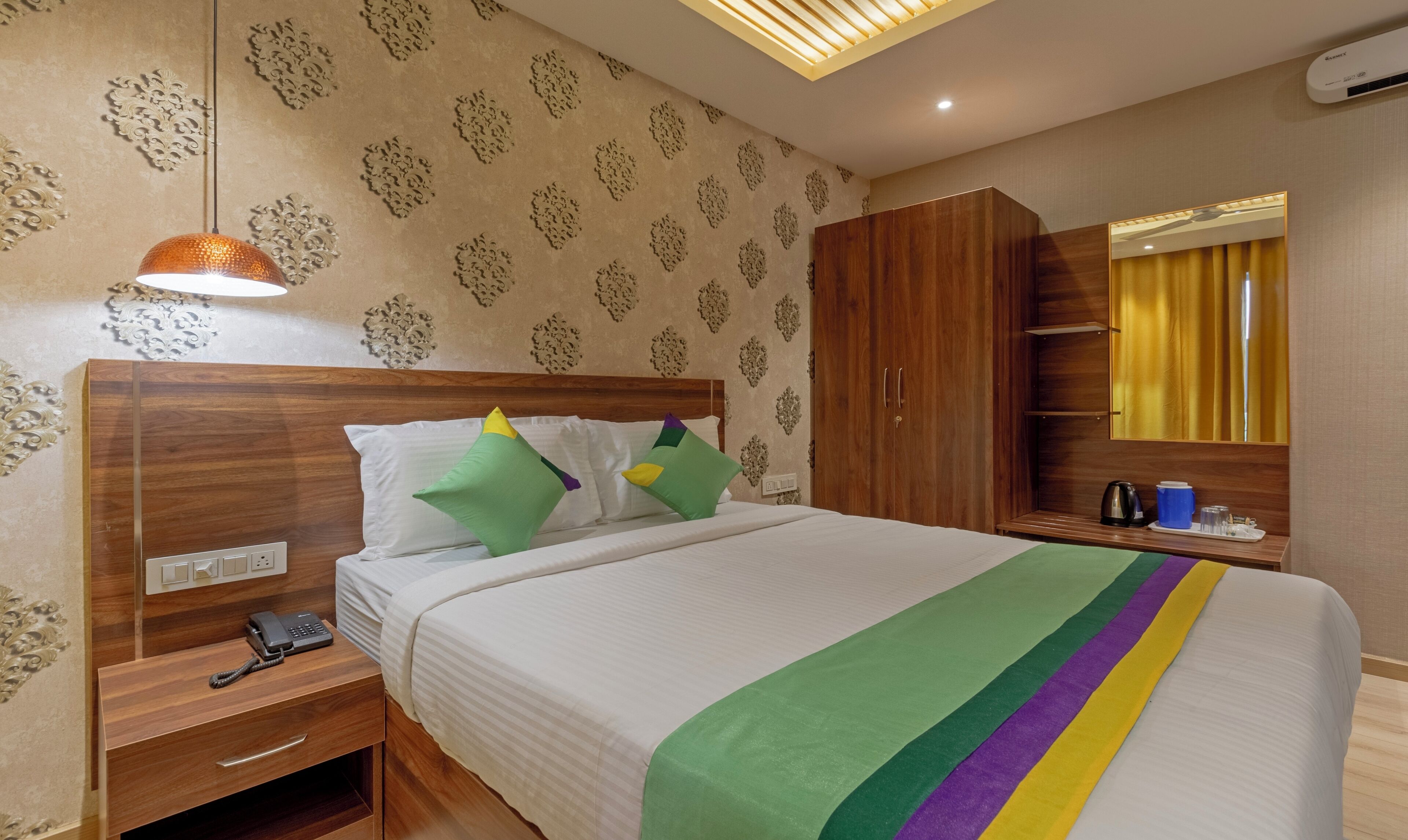 Habitación de hotel con cama, armario de madera, mesita de noche con teléfono, espejo y lámpara en el techo.