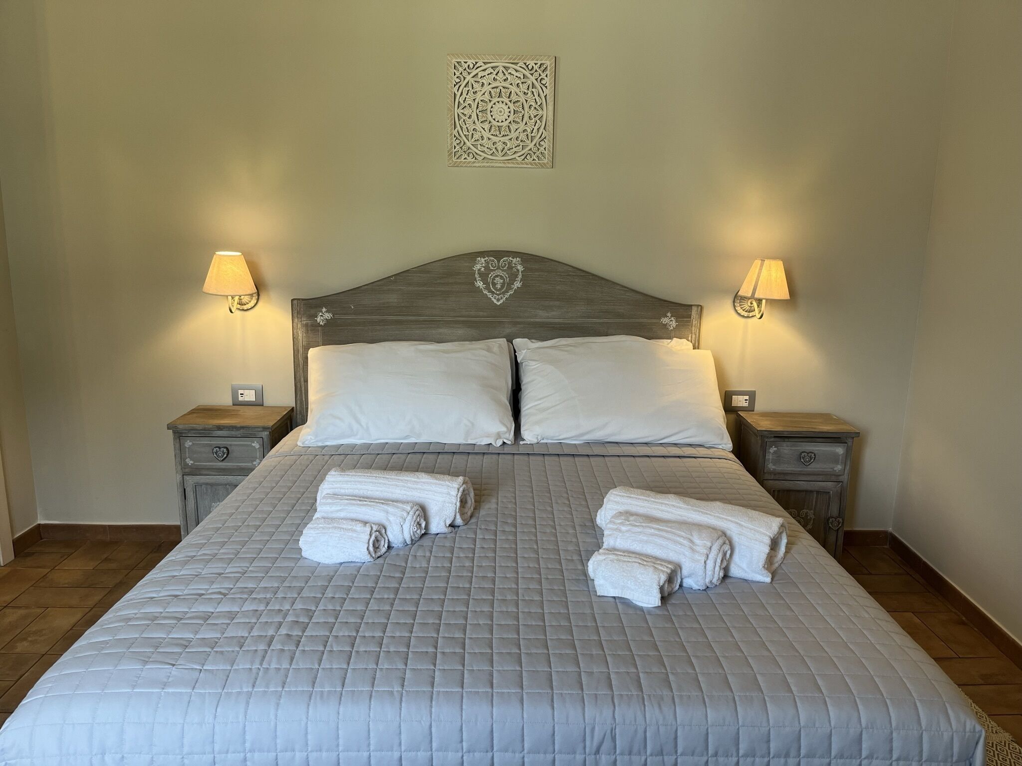 A neatly made bed with a quilted grey bedspread, two white pillows, and two rolled towels on the bed. There are two bedside tables with lamps on either side of the bed.