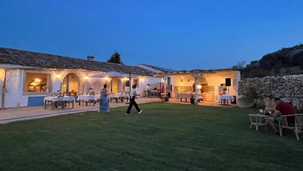 A rustic outdoor dining area with tables and chairs, a stone wall, and a building with arched windows. - Masseria Santacatrini (Melilli)