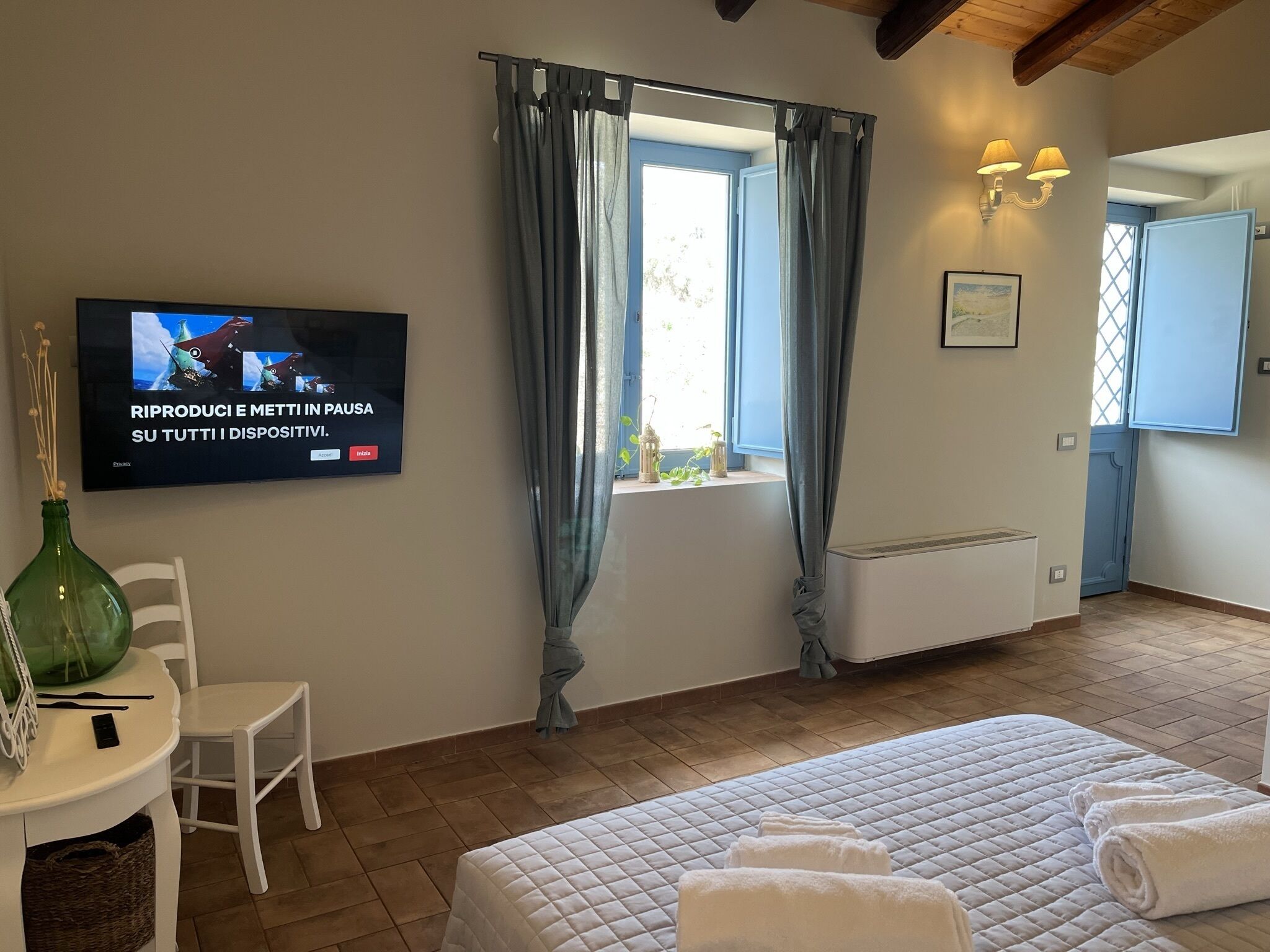 A hotel room with a bed, a TV mounted on the wall, a window with curtains, a small table with a chair, and a bedside table with a vase.