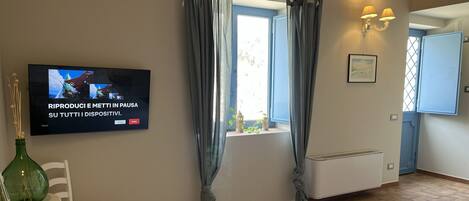 A hotel room with a bed, a TV mounted on the wall, a window with curtains, a small table with a chair, and a bedside table with a vase.