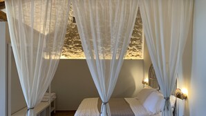 A bedroom with a bed, white curtains, a stone wall, and a bedside table with a lamp. - Masseria Santacatrini (Melilli)