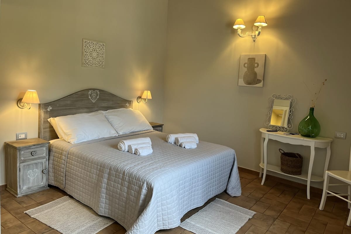 A neatly arranged bedroom with a bed, bedside lamps, a bedside table with a vase, and a mirror.