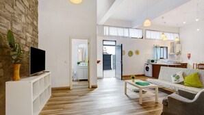 Ett modernt vardagsrum med en platt-TV, trägolv, en soffa, ett soffbord och ett köksområde som syns genom en öppen dörr.