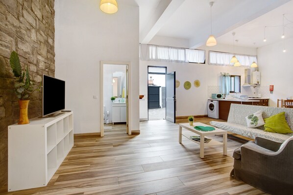 A modern living room with a flat-screen TV, wooden flooring, a sofa, a coffee table, and a kitchen area visible through an open door.