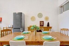 Uma sala de jantar com mesa de madeira posta para quatro pessoas, uma geladeira e uma cafeteira em um armário de madeira.