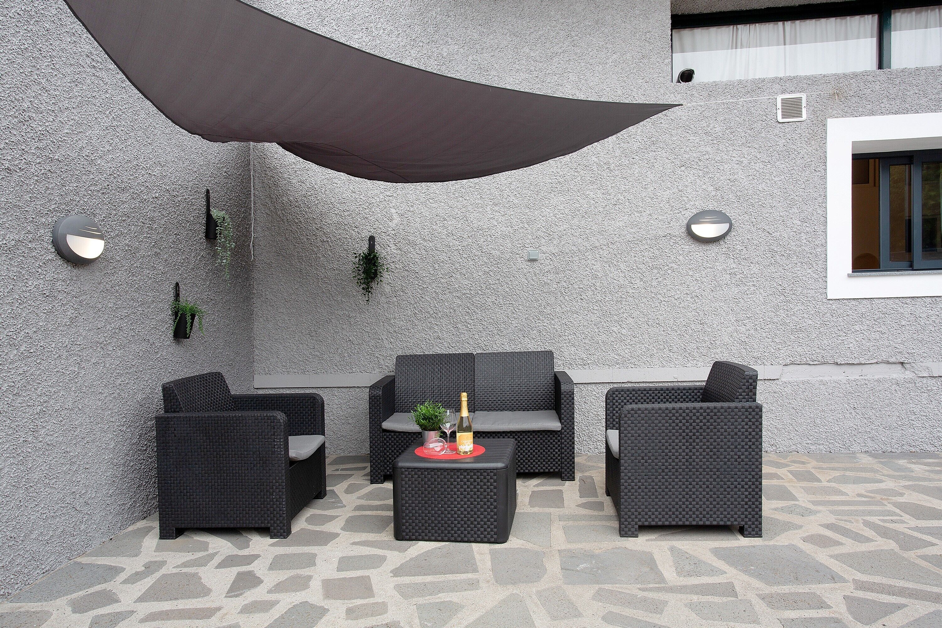 Un espace extérieur aménagé avec des meubles en osier, une table avec une bouteille et une grande voile d’ombrage.