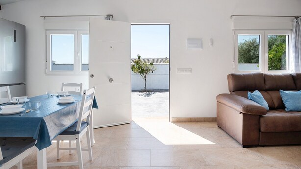 Une pièce comprenant une table à manger dressée pour quatre personnes, un canapé en cuir marron et une porte donnant sur un espace extérieur avec un arbre.