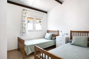 Un dormitorio con dos camas individuales, una ventana con cortinas, una mesita de noche con lámpara y otro mesita auxiliar.