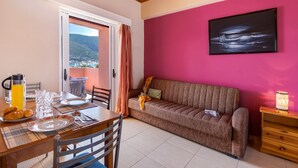 Un coin repas avec une table dressée pour deux personnes, un canapé et une vue sur un paysage montagneux par une porte ouverte.