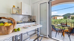 A kitchen with a fruit basket, a dining area with a table and chairs, and a view of buildings and hills through a glass door.
