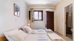 A bedroom with two beds, a window with a curtain, an air conditioning unit, a door with an exit sign, and a view of the bathroom.