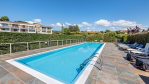 Une piscine rectangulaire, bordée d’une terrasse carrelée, entourée de chaises longues et d’une haie.