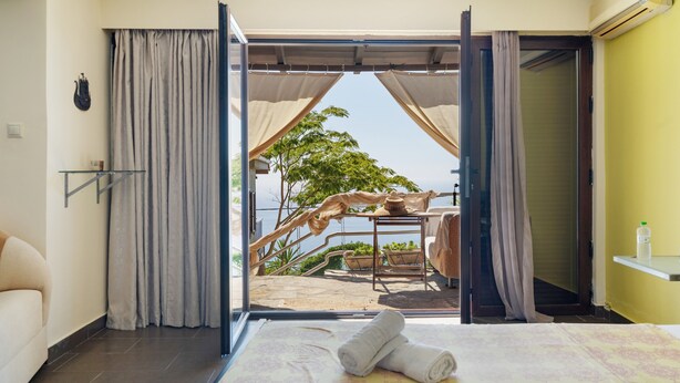 Ein Hotelzimmer mit Bett, Blick auf einen Außenbereich mit Tisch und Stühlen sowie einer Tür zu einem weiteren Raum.