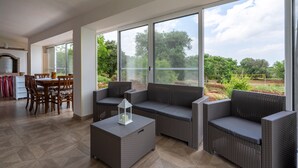 Tilava patio, jossa on rottinkihuonekaluja, ruokailutila puutuoleineen sekä suuret ikkunat, joista avautuu näkymä vehreään maisemaan.