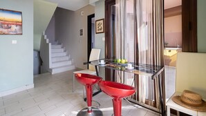 A dining area with a glass table, two red bar stools, a staircase, and a door with curtains.