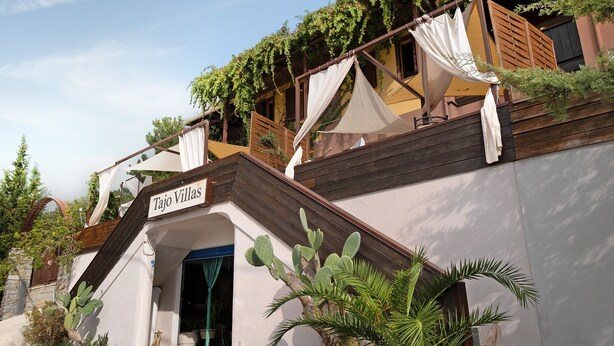 A building named 'Tajo Villas' with a staircase leading to a covered entrance, surrounded by cacti and palm plants.