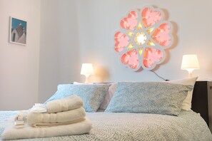 Uma cama bem arrumada com colcha estampada, duas luminárias de cabeceira, uma luminária decorativa na parede e uma pilha de toalhas dobradas.