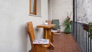 A wooden bench with a cushion, a small table with a white object, a chair with a patterned cushion, and a potted plant with red flowers on a brick patio.