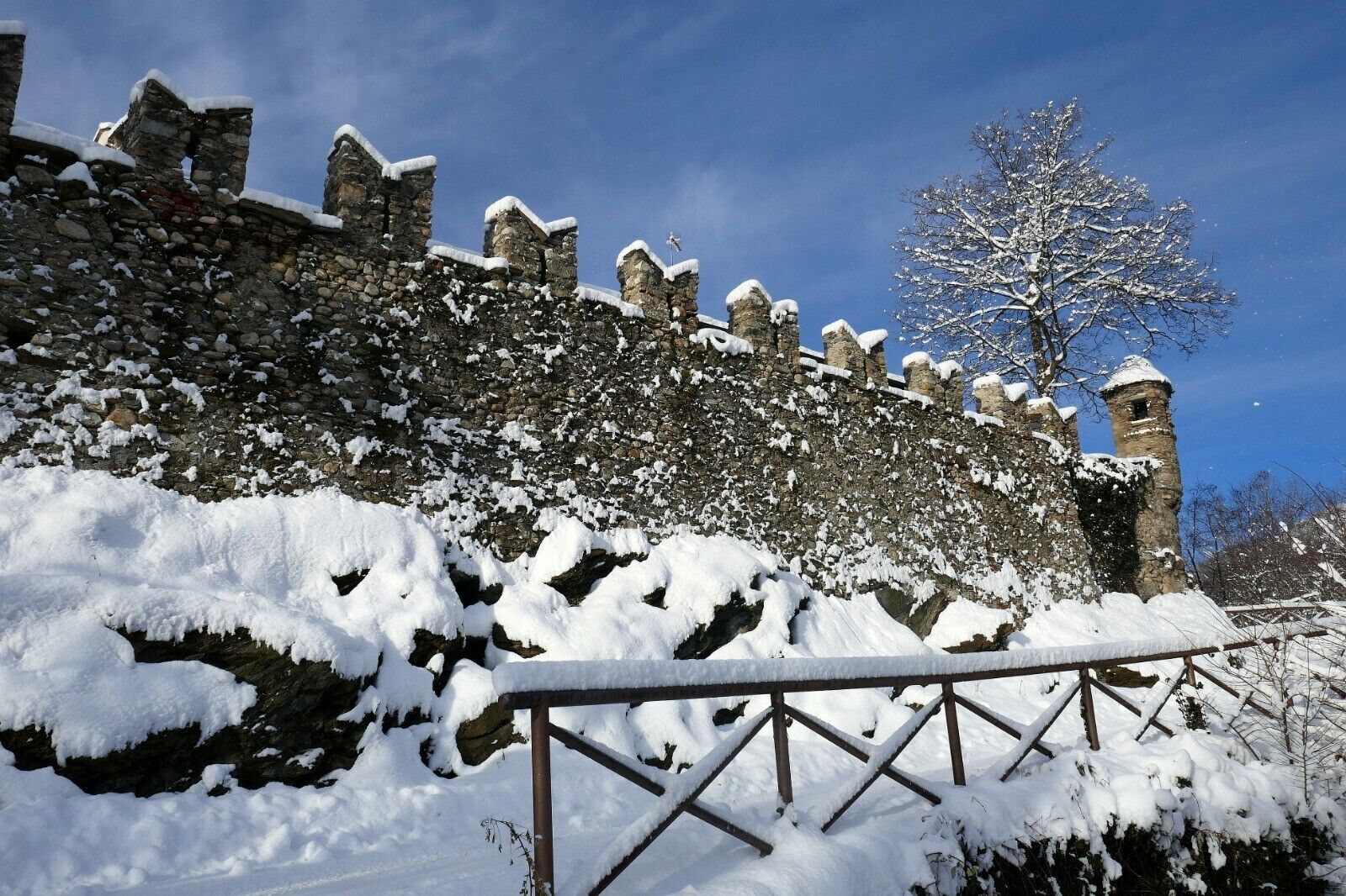 A stone wall with snow accumulation, a metal railing, and a clear blue sky.