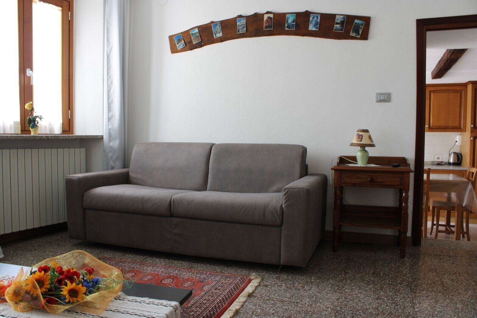 Ein Wohnzimmer mit grauem Sofa, einem kleinen Tisch mit Lampe, einer Wandcollage aus Fotos und einem Essbereich mit Tisch und Stühlen.