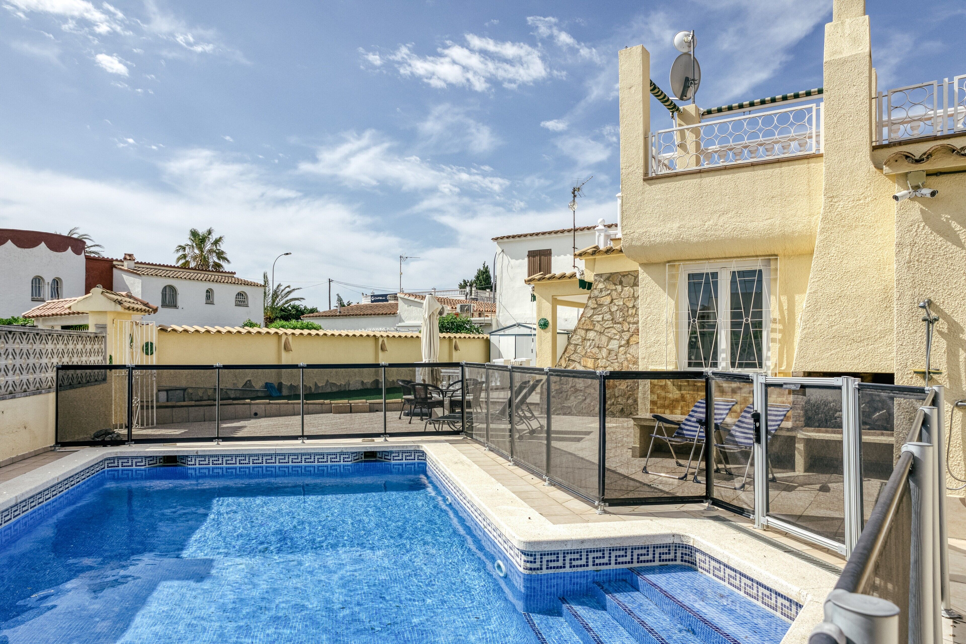 Piscina con borde de azulejos, un porche cubierto con sillas y vistas a casas vecinas.
